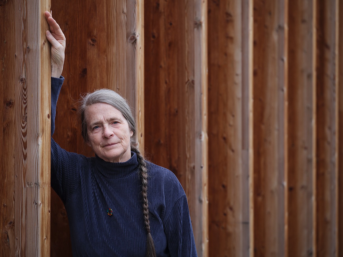 Auf dem Bild ist die Meteorologin und Klimaforscherin Helga Kromp-Kolb zu sehen. Sie trägt einen dunkelblauen Rollkragenpullover und lehnt an einer Holzwand. Ihre Haare sind grau und zu einem langen Zopf geflochten, der ihr über die linke Schulter hängt.